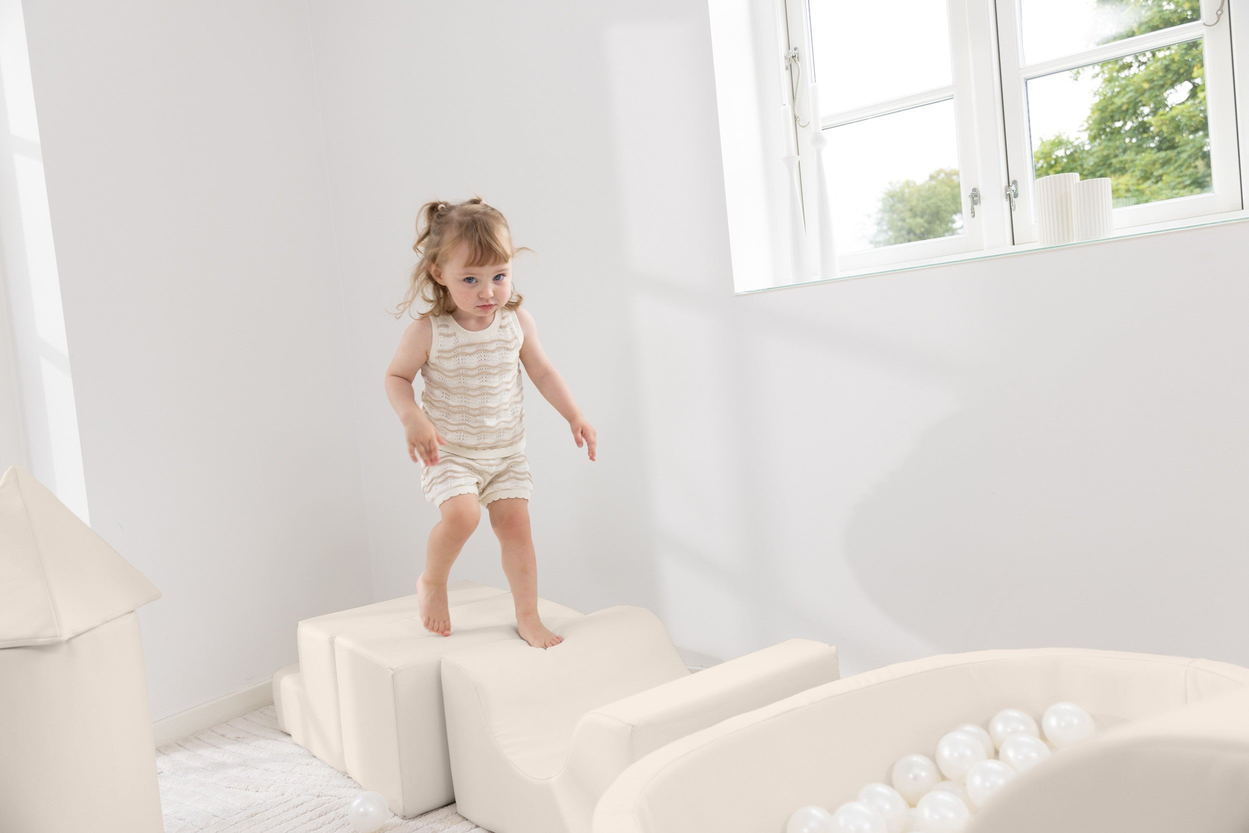 Child standing on foam blocks in a bright, minimalistic room with large windows.