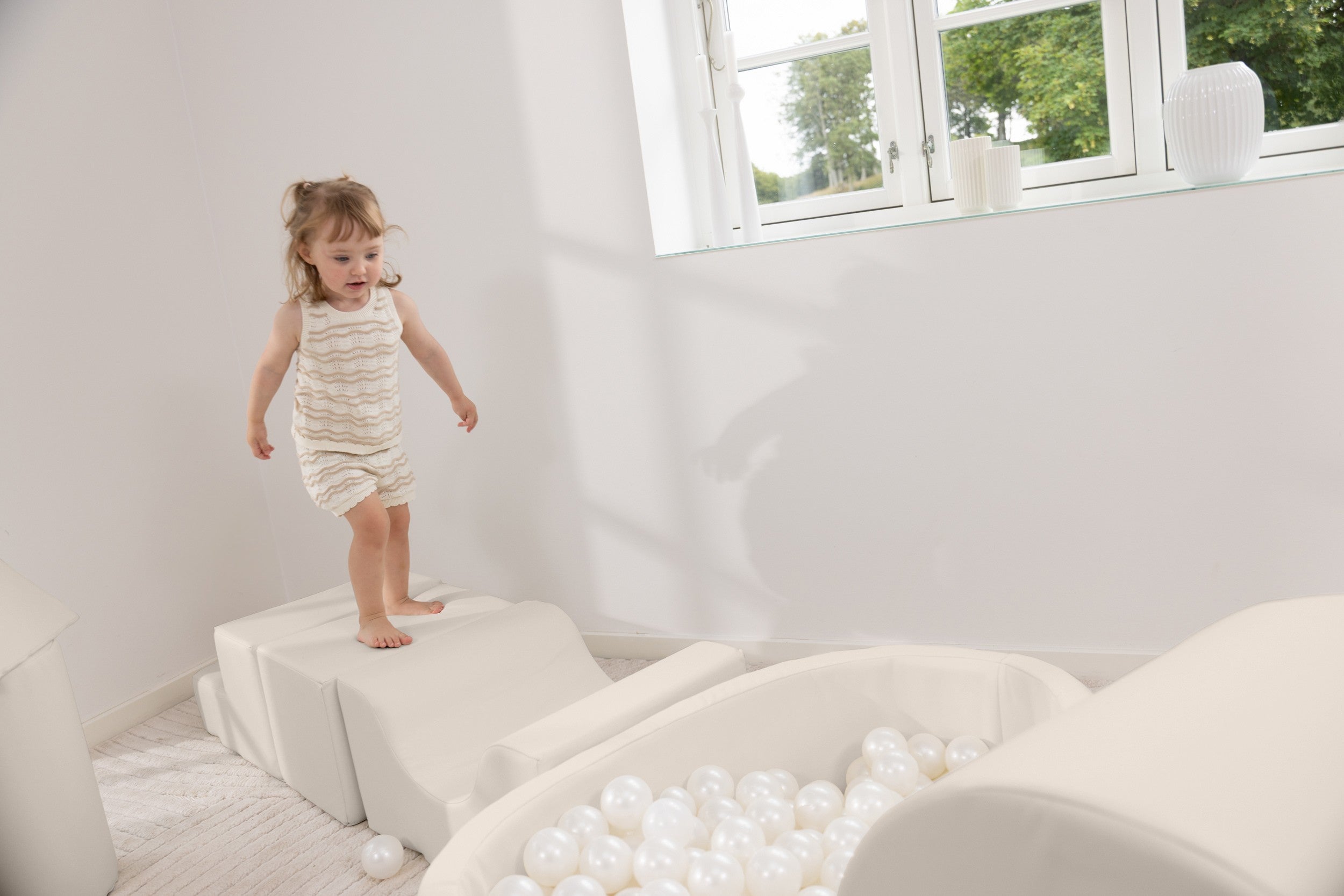 Child standing on a cushion in a room with a window and ball pit.